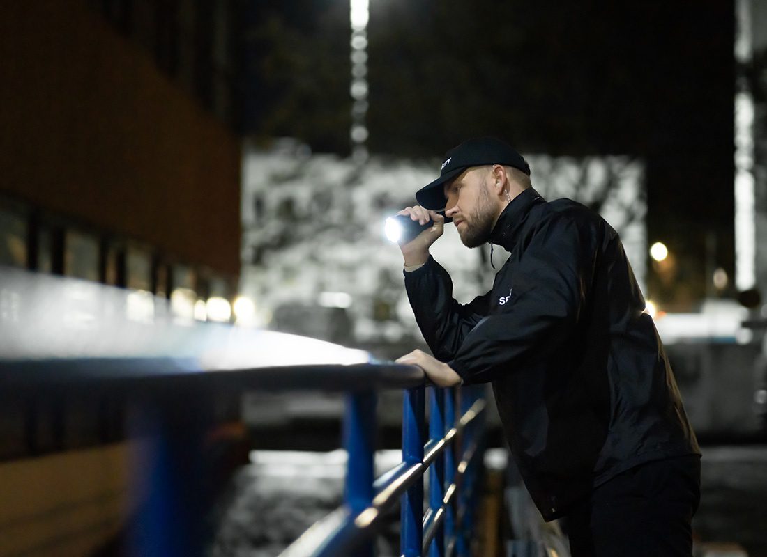 Insurance by Industry - Portrait of a Male Security Guard Pointing a Flash Light Outside Next to a Commercial Building to Investigate Suspicious Activity