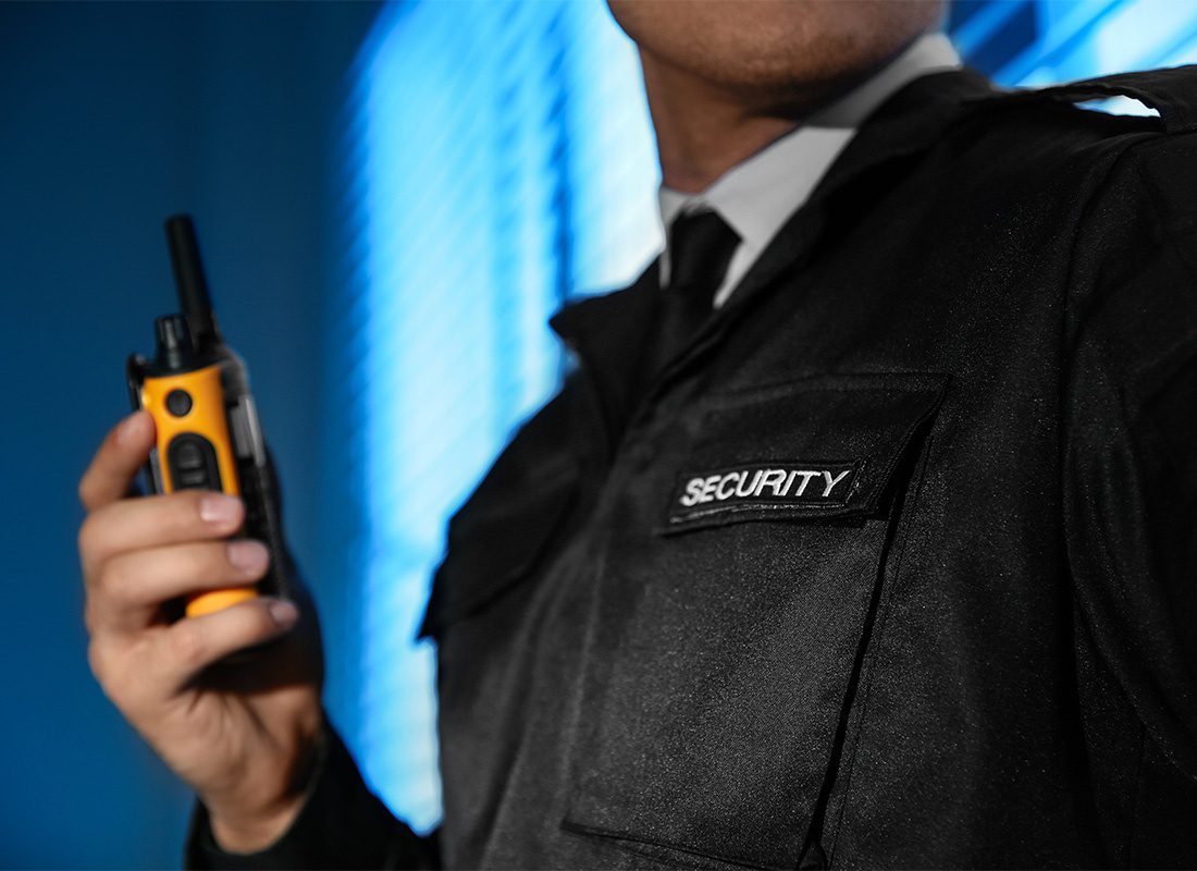 Security Guard Insurance - Closeup View of a Male Security Guard Wearing a Uniform Standing in a Blue Lit Room While Holding a Walkie Talkie in his Hand